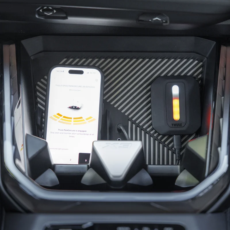 Interior view of a car showing the Thule Epos ParkSecure sensor unit and a smartphone displaying the app's parking assistance interface.