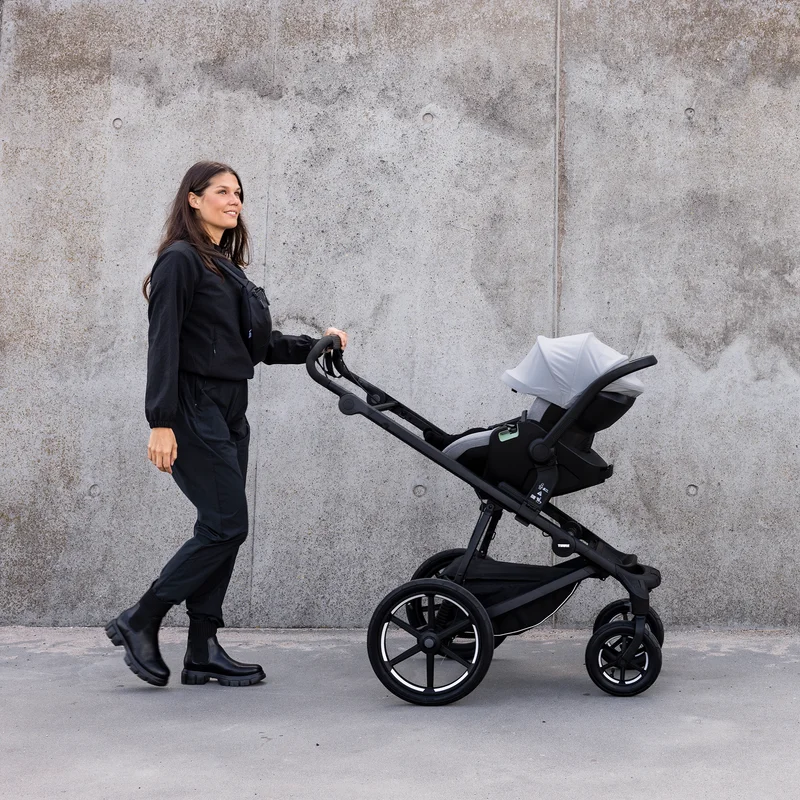 Eine Frau, die in Schwarz gekleidet ist, schiebt einen Thule Urban Glide 4-Rad-Kinderwagen mit einem Thule Maple Babysitz in hellgrau darauf vor einer großen Betonwand.