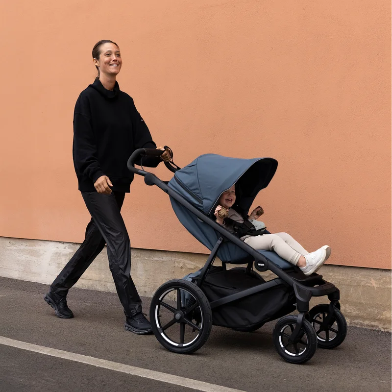 Frau schiebt Thule Urban Glide 4-Rad-Kinderwagen mit Kindersitz an Betonwand entlang.