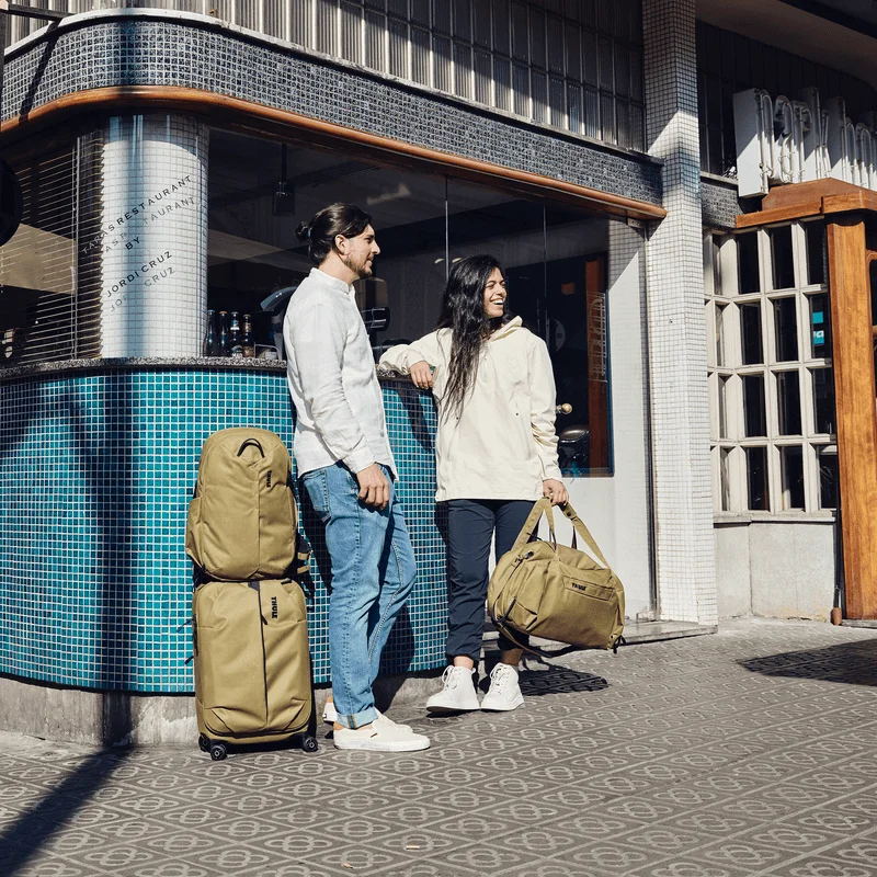 Ein Paar steht mit hellbraunen Taschen in der Sonne neben einem Restaurant, unter den Taschen auch ein Thule Aion Handgepäckkoffer