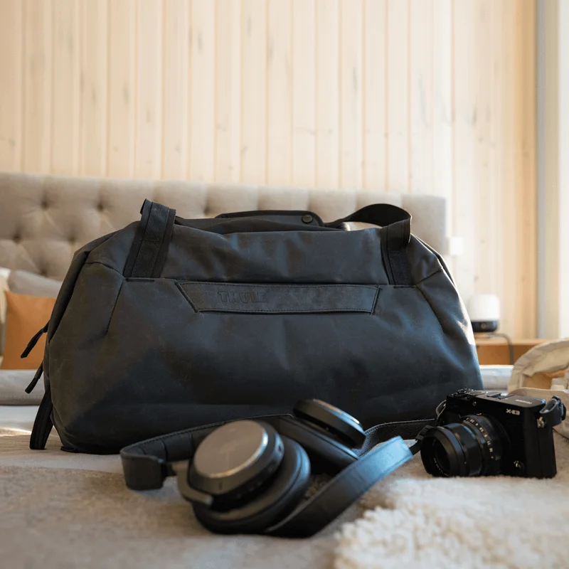 Eine schwarze Thule Aion Reisetasche liegt auf einem Bett mit Kopfhörern und einer Kamera in einem Hotelzimmer.