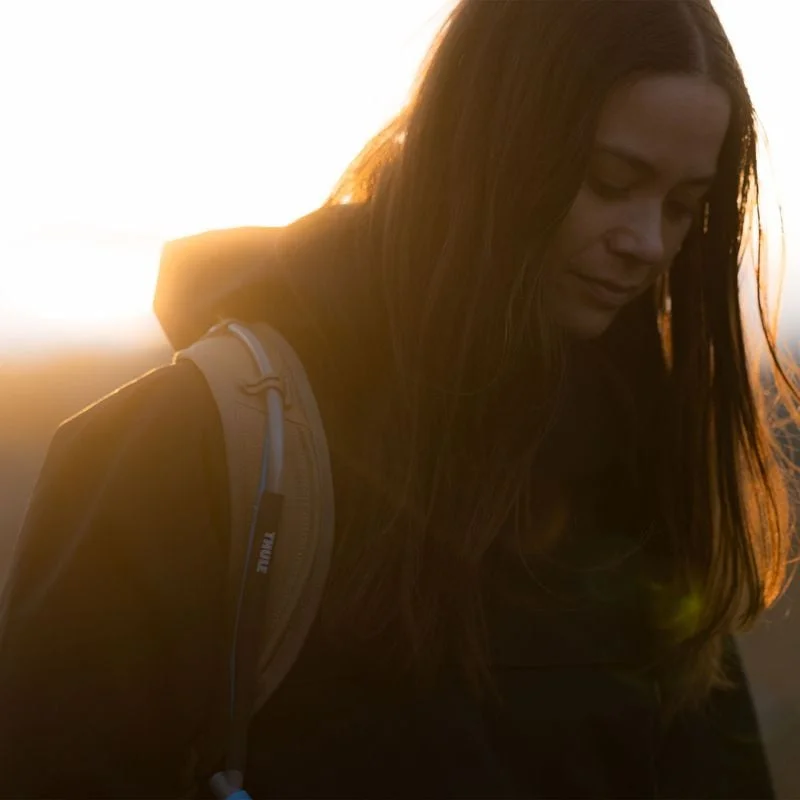 A woman stands in a black rain jacket in the sunshine carrying a beige Thule AllTrail hydration backpack.