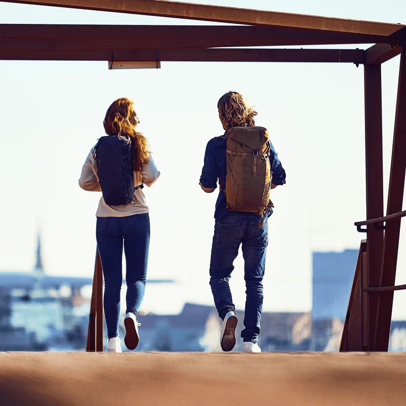 A man and women walk down a street carrying black and tan Thule AllTrail X hiking backpacks.