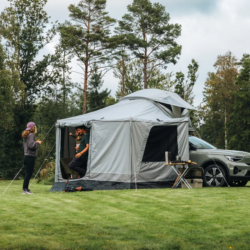 Thule Approach 2 soft-shell rooftop tent unfolded and mounted with awning and walls on the top of a car parked in an outdoor setting and a family interacting in the tent.
