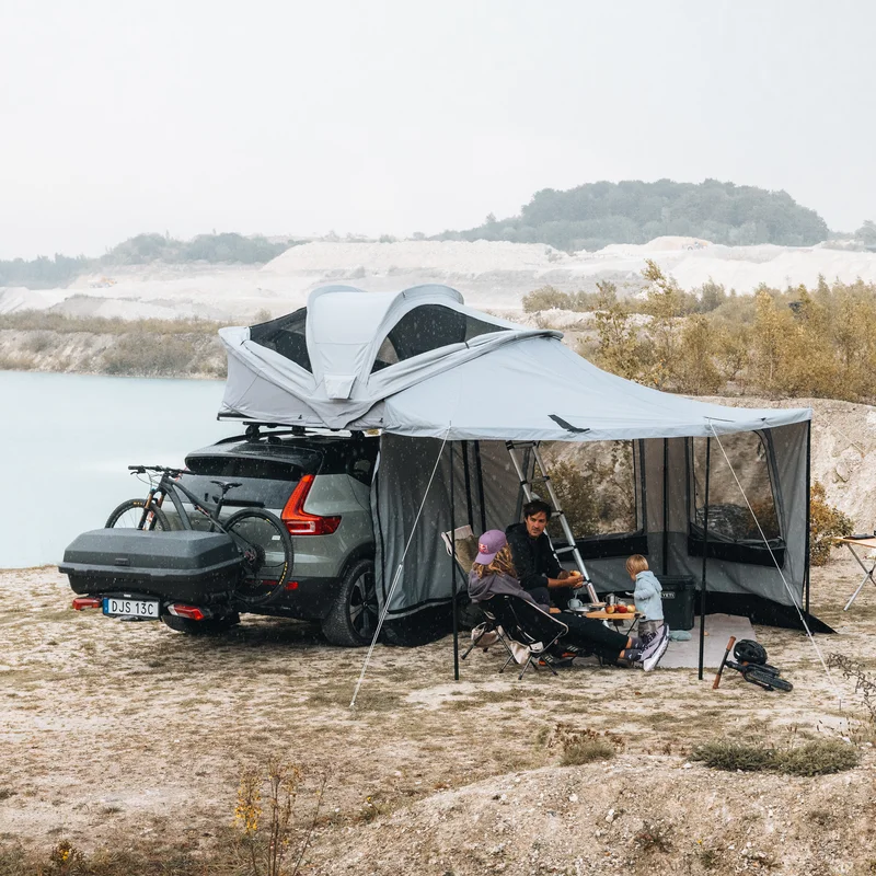 Thule Approach 2 soft-shell rooftop tent unfolded and mounted with awning and walls on the top of a car with one bike and Thule Santu cargo box on the towbar bike rack parked in an outdoor setting and a family interacting by the tent.