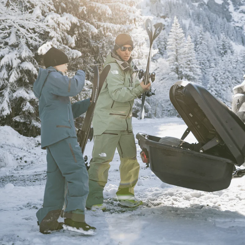 Two people dressed in ski gear, prepare to load their ski equipment into an open, Thule Arcos XL, surrounded by snow-covered trees and winter sunshine.