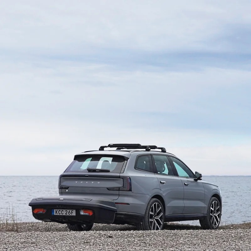A dark grey car with Thule Arcos XL hard-shell rear cargo carrier mounted on the back, is parked by a shoreline, with roof racks on the top of the car.