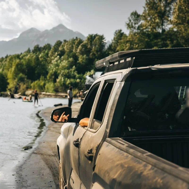 A man is sitting in a off-road pickup truck with a Thule Caprock roof platform and a rail mount on the roof looking at some friends doing activities at a nice outdoors setting with a river.
