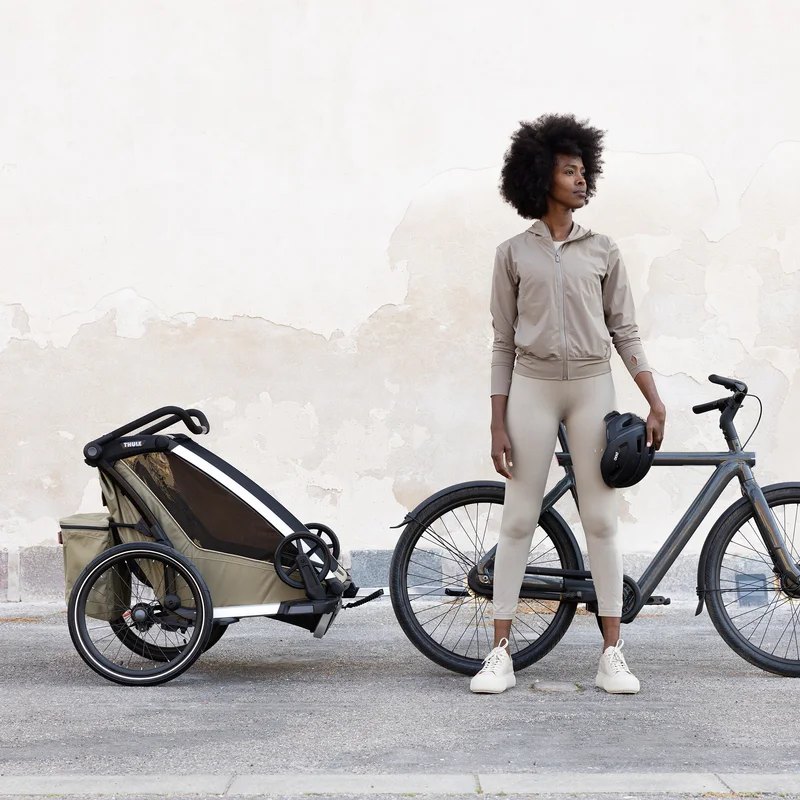 Eine Frau steht mit einem Fahrrad und einem braunen Thule Chariot Kinderfahrradanhänger neben einer Betonwand .