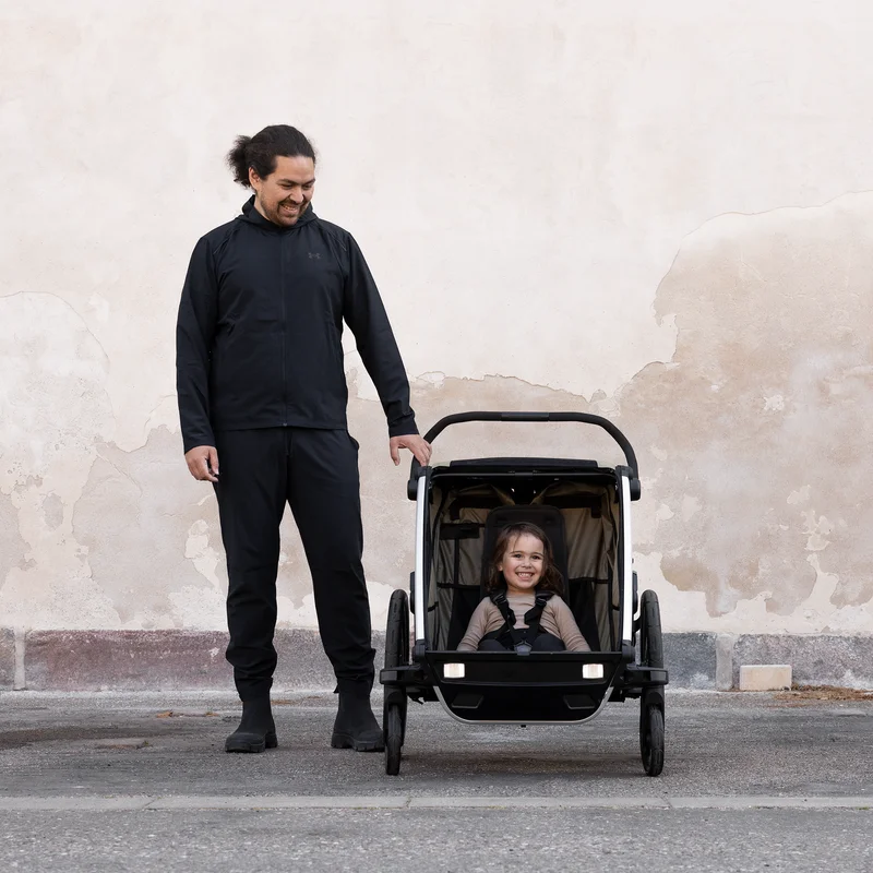 Ein Mann steht neben einem braunen Thule Chariot Kinderfahrradanhänger und ist komplett in schwarz gekleidet.