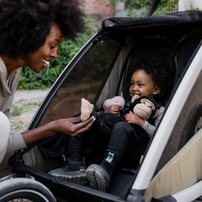 Lächelndes Kind sitzt in einem Thule Chariot Cross 2 Fahrradanhänger und Kinderwagen, während das Elternteil sich hineinlehnt und beim Spaziergang lächelt.