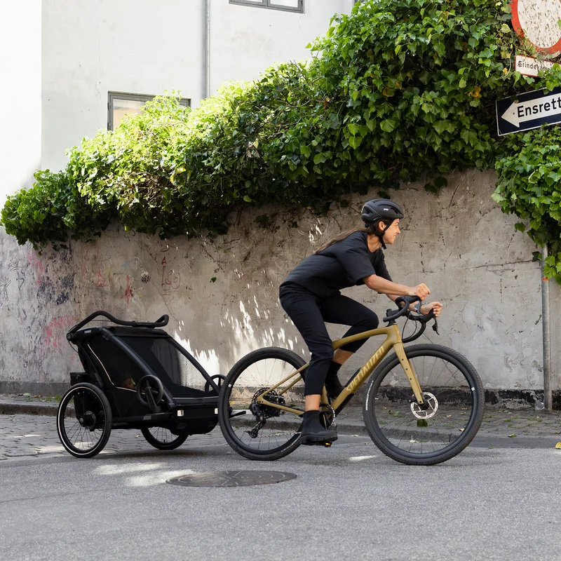 Eine Frau fährt mit ihrem schwarzen Thule Chariot Sport 2 Kinderfahrradanhänger eine sonnige Straße hinunter.