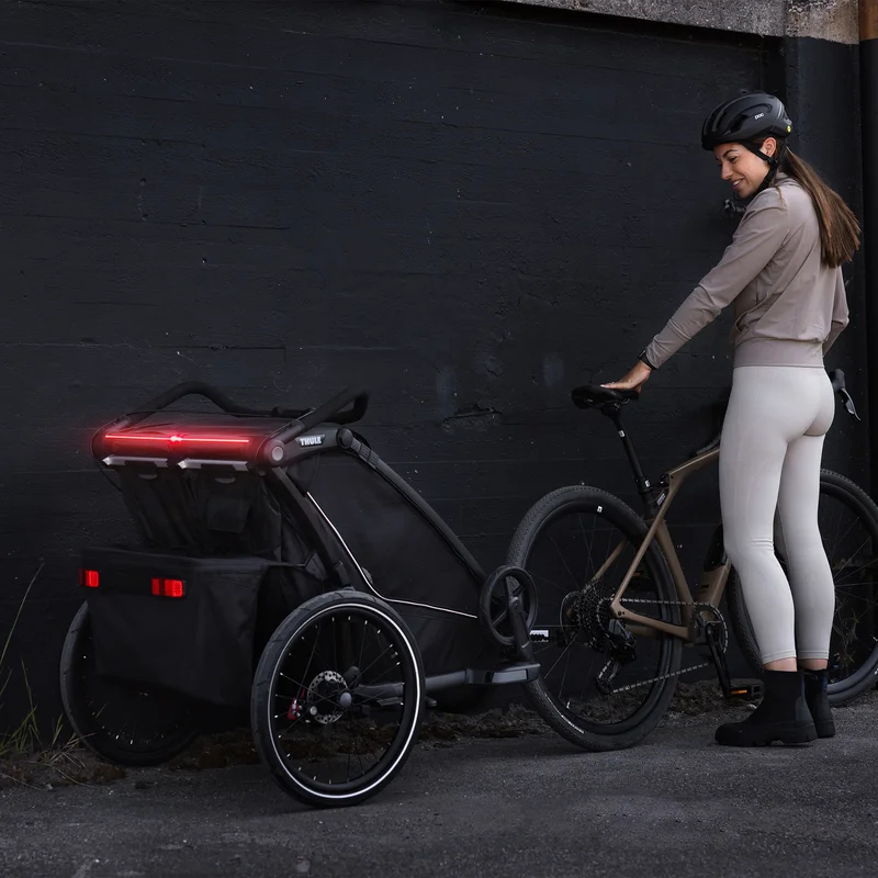 Eine Frau blickt auf ihren schwarzen Thule Chariot Sport 2 Kinderfahrradanhänger mit integrierten LED-Leuchten zurück.
