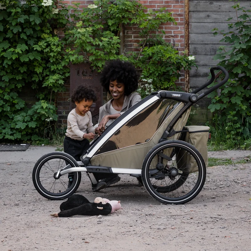 Eltern mit Kind neben einem Thule Chariot Fahrradanhänger mit Geländeset, draußen auf einem Kiesweg geparkt.