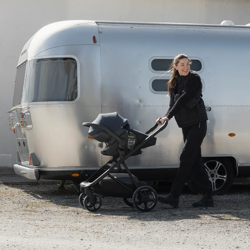 Adult pushing a Thule Charm stroller with a Thule Maple infant car seat attached to it beside a camper trailer outdoors.