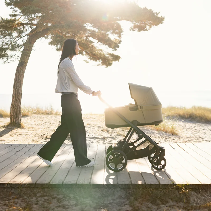 Eine Frau schiebt einen Thule Charm Kinderwagen eine sonnige Strandpromenade entlang.