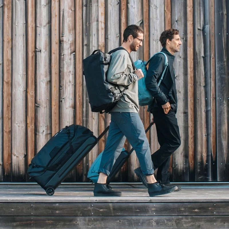 Two men are walking down a sunny path with black and blue Thule Chasm duffels, backpacks and suitcases.