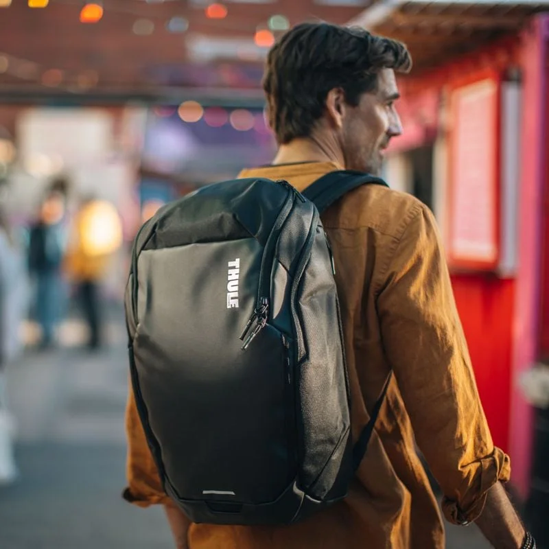 A man in an orange shirt is carrying a black Thule Chasm backpack.