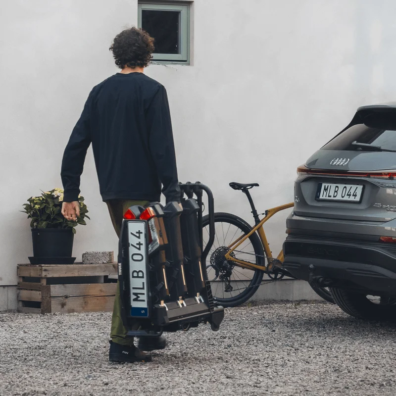 A man in casual outdoor clothing carries a bike rack by its handle, walking between a house and an SUV parked on a gravel driveway.