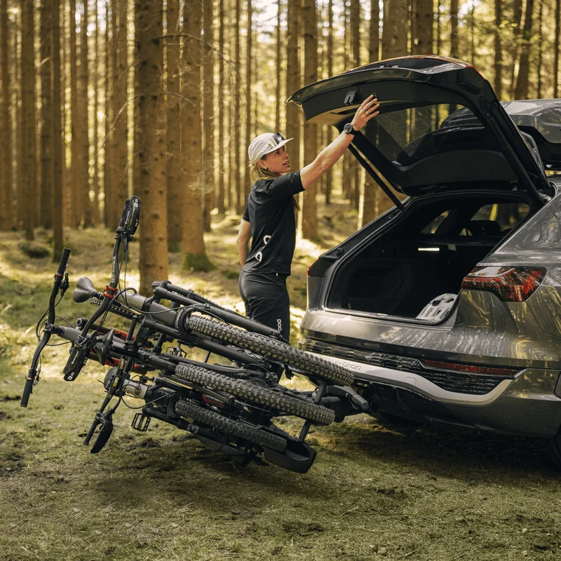 A woman in a forest has tilted her bike rack and opens the trunk of an SUV, preparing to load or unload her bike gear.