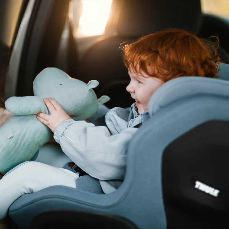 Ein orangehaariges Kind lächelt und hält ein Stofftier, während es in einem Thule Elm Kleinkinderautositz sitzt.