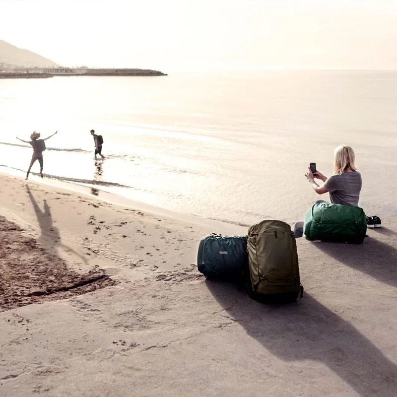 A woman sits next to the beach with Thule Landmark travel backpacks around her and she is taking a picture of her friends.