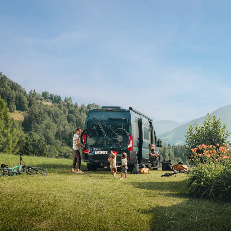 A camper van set up in a scenic outdoor location, featuring the Thule VeloTrack with the Thule Lightboard Van. The bikes are mounted at a lower position, ensuring taillights remain visible while making loading easier.