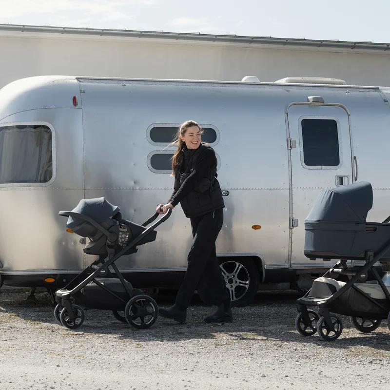 Adult pushing a stroller with a Thule Maple Connect infant car seat attached to it beside a camper trailer outdoors.