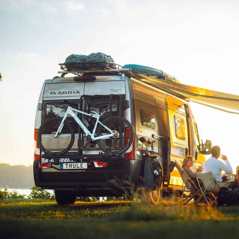 A camper van parked near a scenic lakeside location at sunset, equipped with the Thule VeloTrack bike rack carrying a mountain bike. Two people relax under the extended awning, enjoying the outdoor setting.
