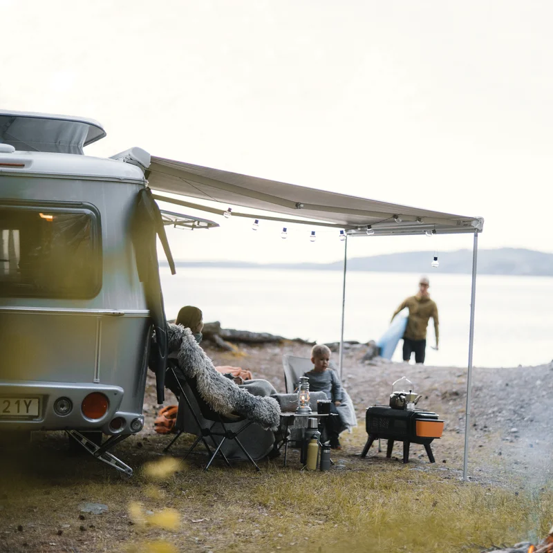 A man with a surfboard walks towards a woman sitting by a white van with a Thule Omnistor 6300 van awning.