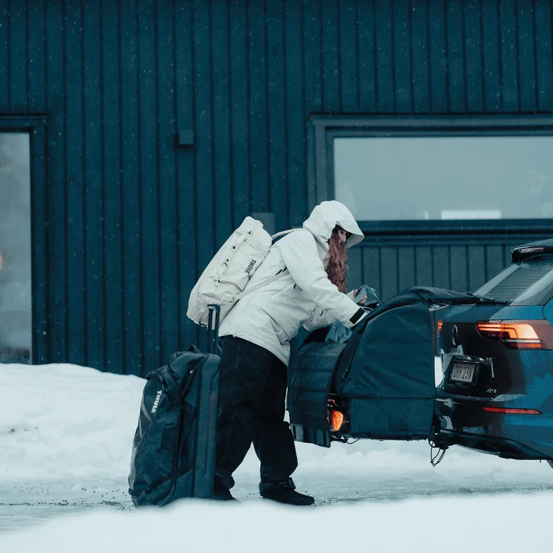 A person in a winter coat loads a large bag into the Thule Onto 2, a towbar-mounted cargo bag, attached to the rear of a blue station wagon in a snowy environment.