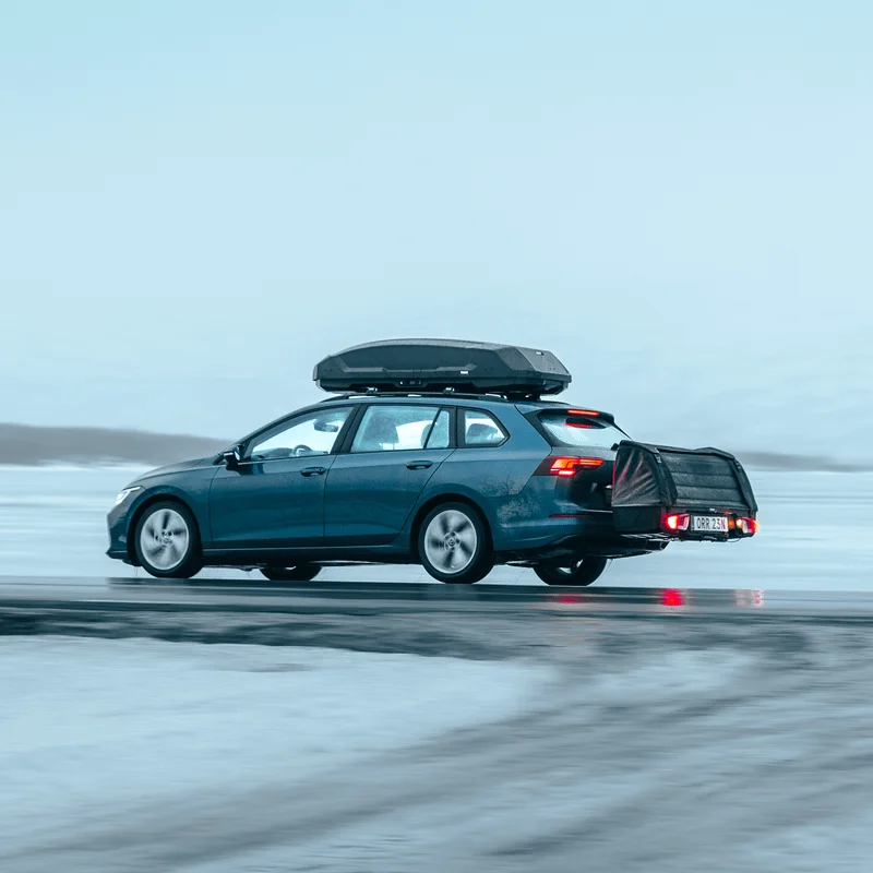 A blue station wagon drives through a snowy landscape with the Thule Onto 2 cargo bag mounted on the towbar and a Thule Force 3 roof box on top, carrying extra gear for winter travel.