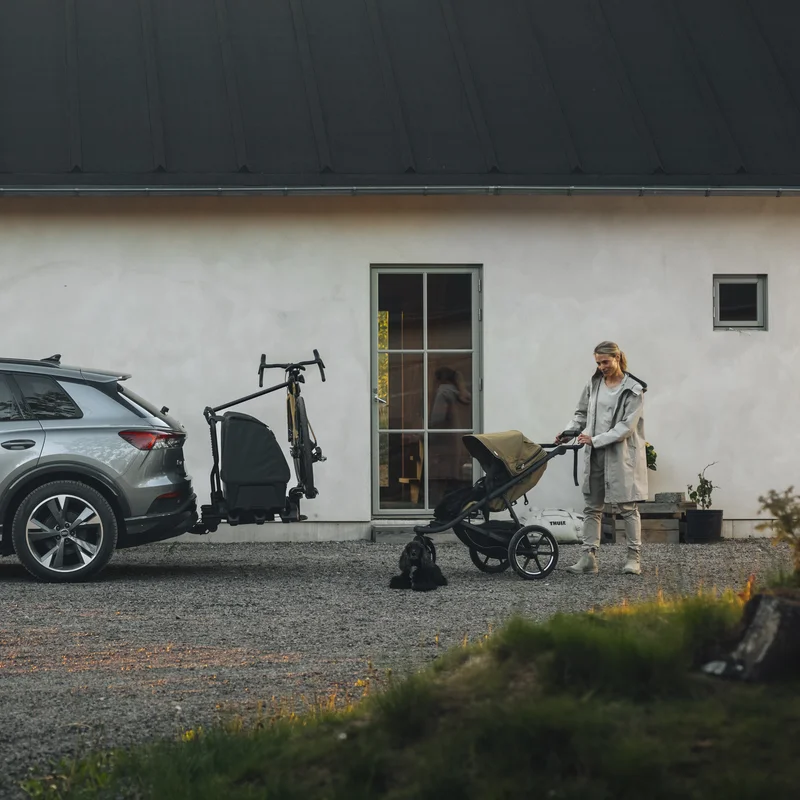 A woman is pushing a Thule Urban Glide 3 stroller next to a car with a Thule Onto 2 towbar cargo carrier on a Thule EasyFold 3 towbar bike rack with a bike arm holding a bike.