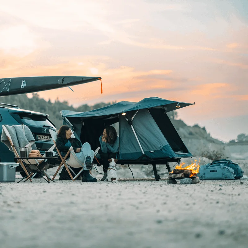A woman is sitting inside a Thle Outset car tent next to a car, talking to another woman in an outdoors summer camping environment.