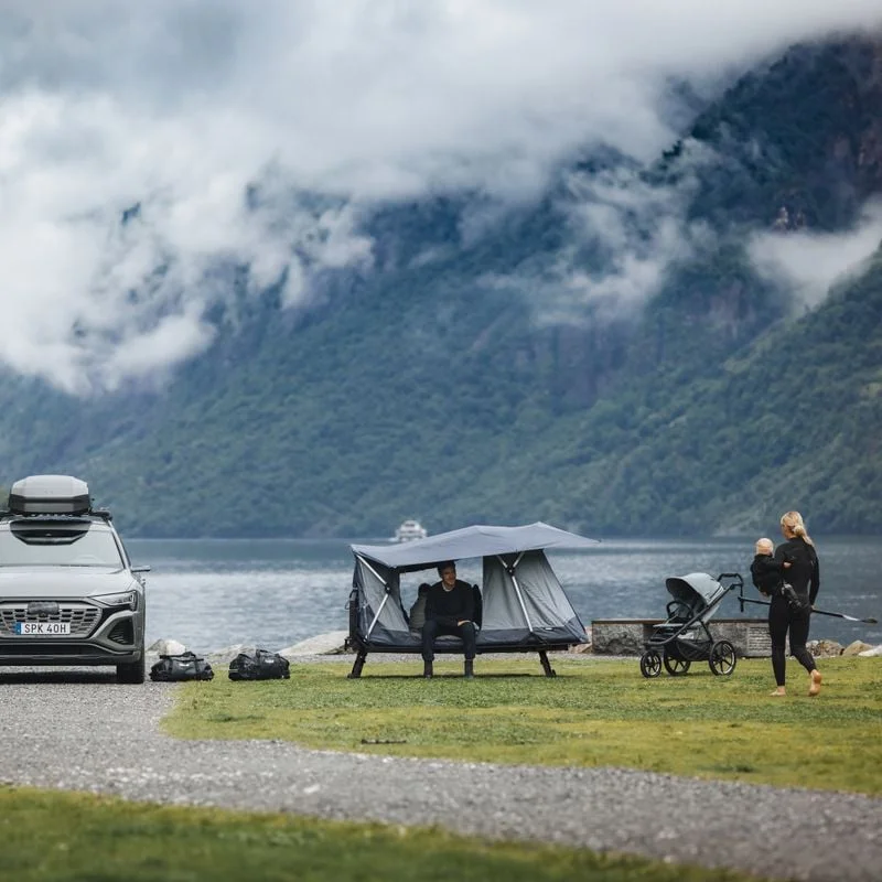 A serene lakeside campsite featuring a Thule Outset tent set up beside a parked SUV with a roof box. A person sits under the tent's shade, while another walks nearby with a stroller.