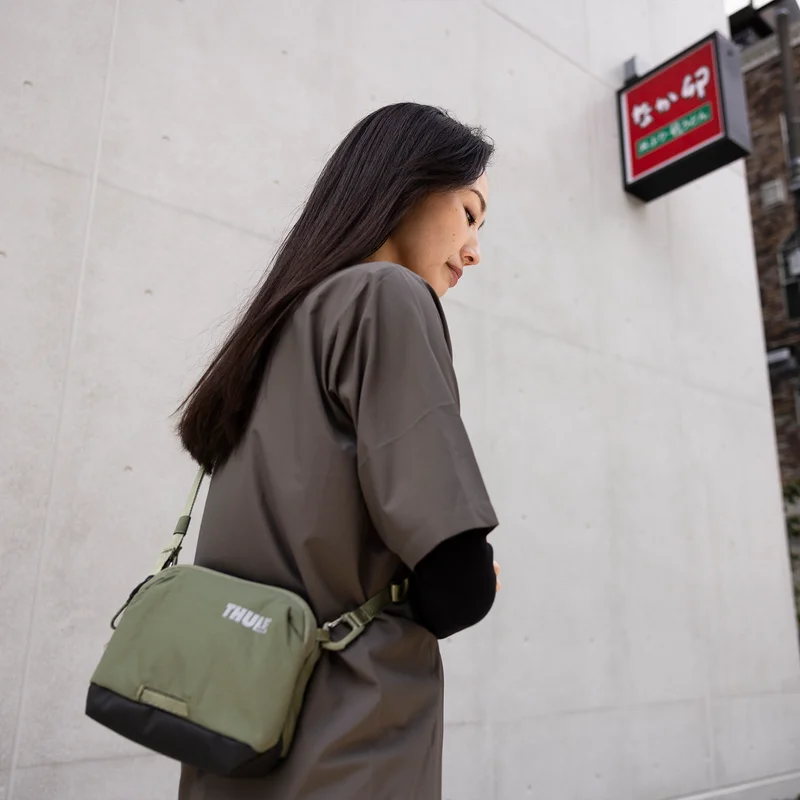 A woman stands against white wall with a Thule Paramount 2L crossbody bag.