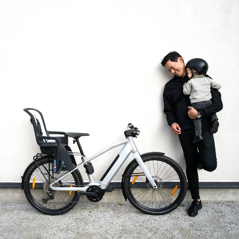 Ein Mann lehnt mit seinem Kind und einem Fahrrad mit einem Thule RideAlong2 Fahrradkindersitz an einer Wand .