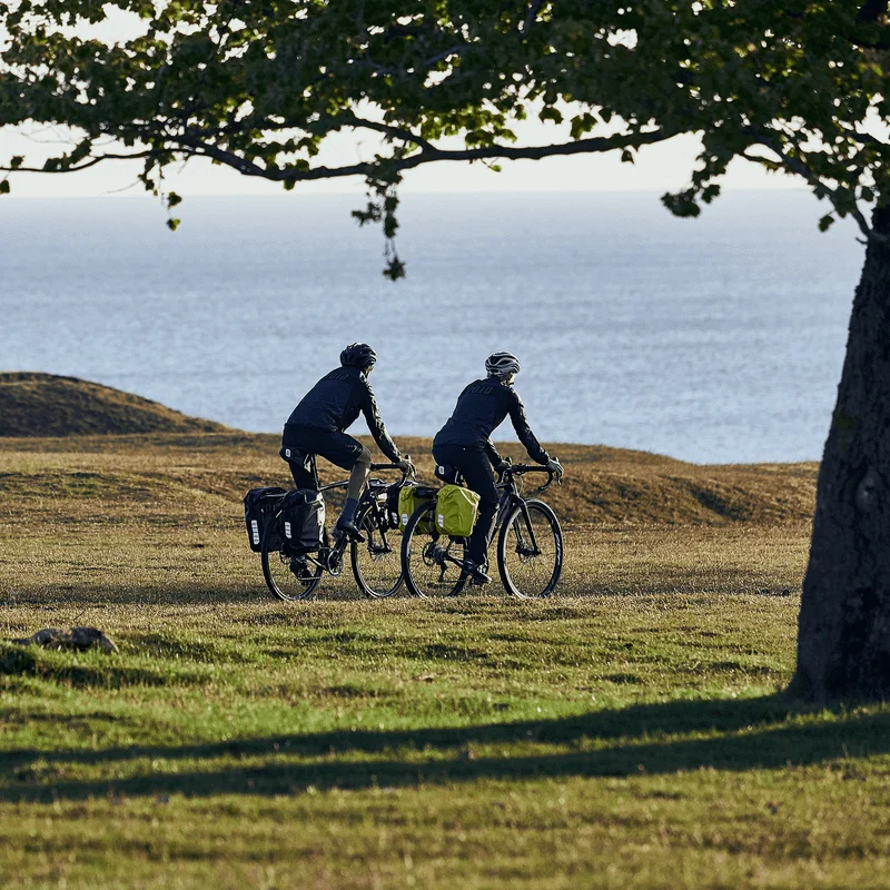 A couple bike by the ocean, with bikes that have black and yellow Thule Shield 25L bike panniers.