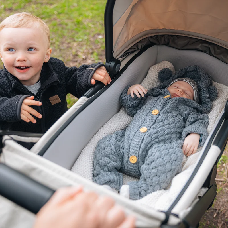 Baby liegt im Thule Spring 2 Kinderbett, während das Geschwisterkind in der Nähe lächelt.