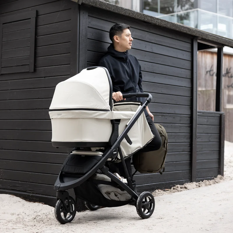 A man holding the bar of the Thule Spring 2 stroller with a light beige bassinet in front of a black wooden cabin.