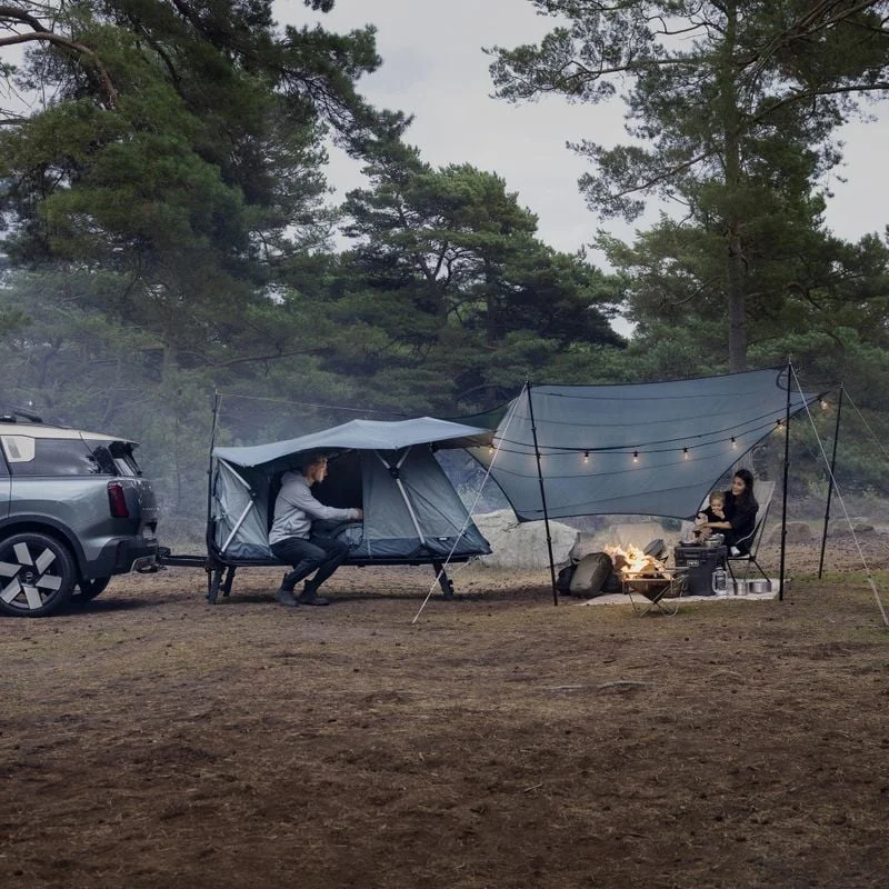 A Thule Outset tent set up next to a parked car in a forest. A Thule Starlet tarp provides additional coverage over the entrance, creating a cozy outdoor space. A person is seated near a campfire, enjoying the natural surroundings.