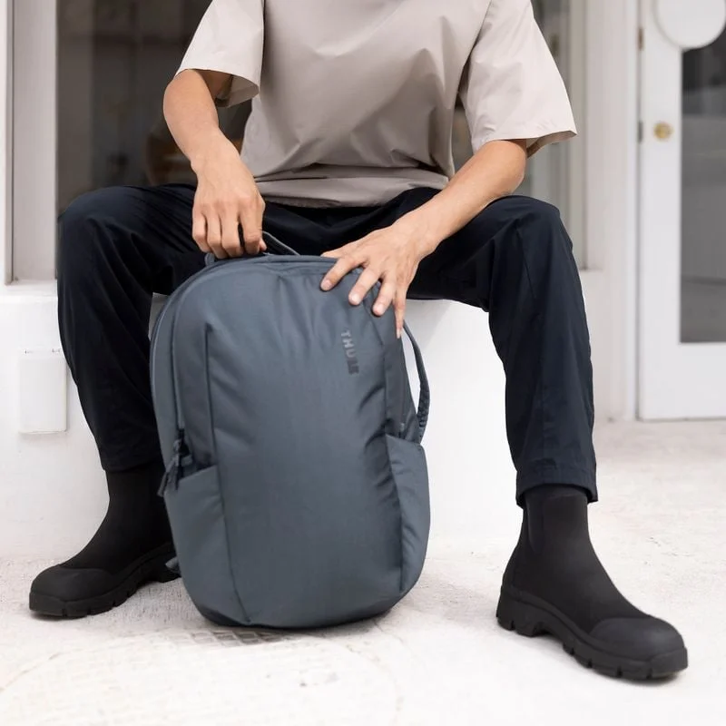 A close-up of someone opening a blue Thule Subterra backpack while sitting next to a building.