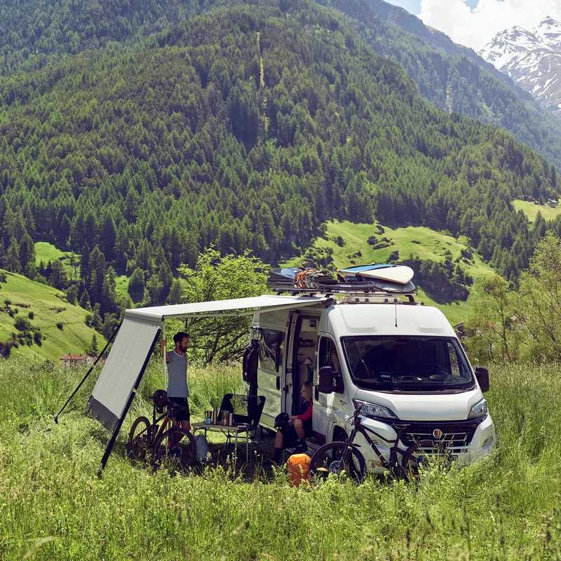 A white van is parked in the grass with a white Thule Sun Blocker Front awning side wall.