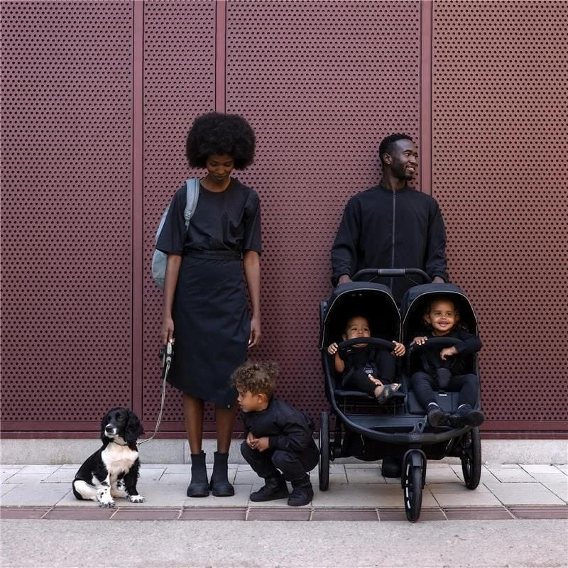 A man, woman, and child walking in front of a dark red wall with the Thule Urban Glide 3 double stroller. One child is seated inside the stroller.