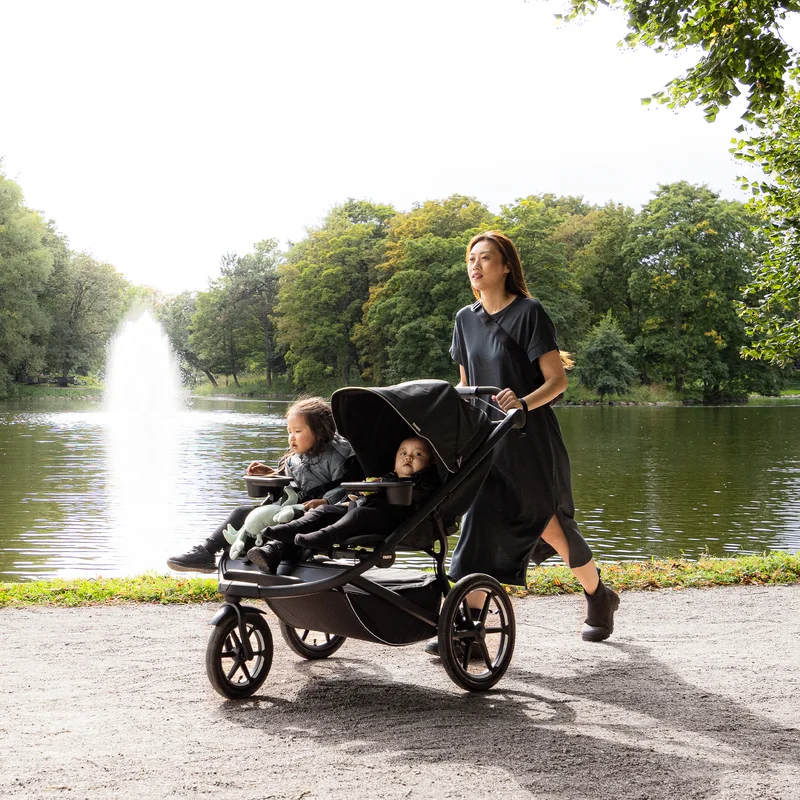 A woman walking through the park with two children seated in the Thule Urban Glide 3 double stroller.