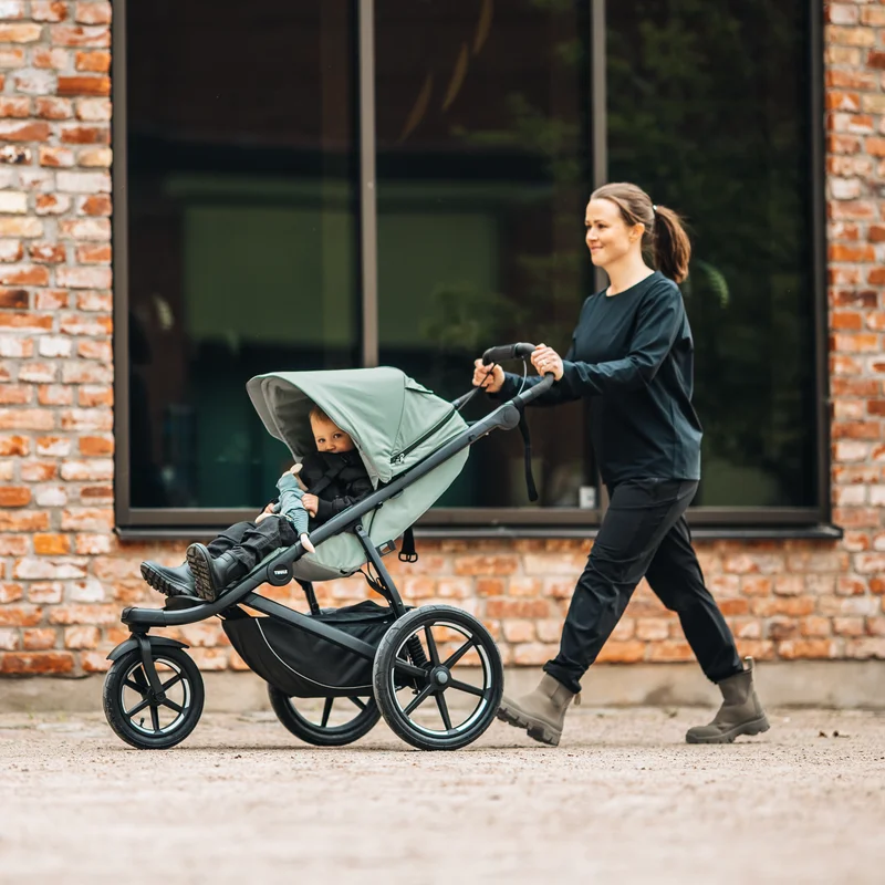 Frau geht mit dem Thule Urban Glide 3 Kinderwagen an einer Ziegelmauer vorbei.