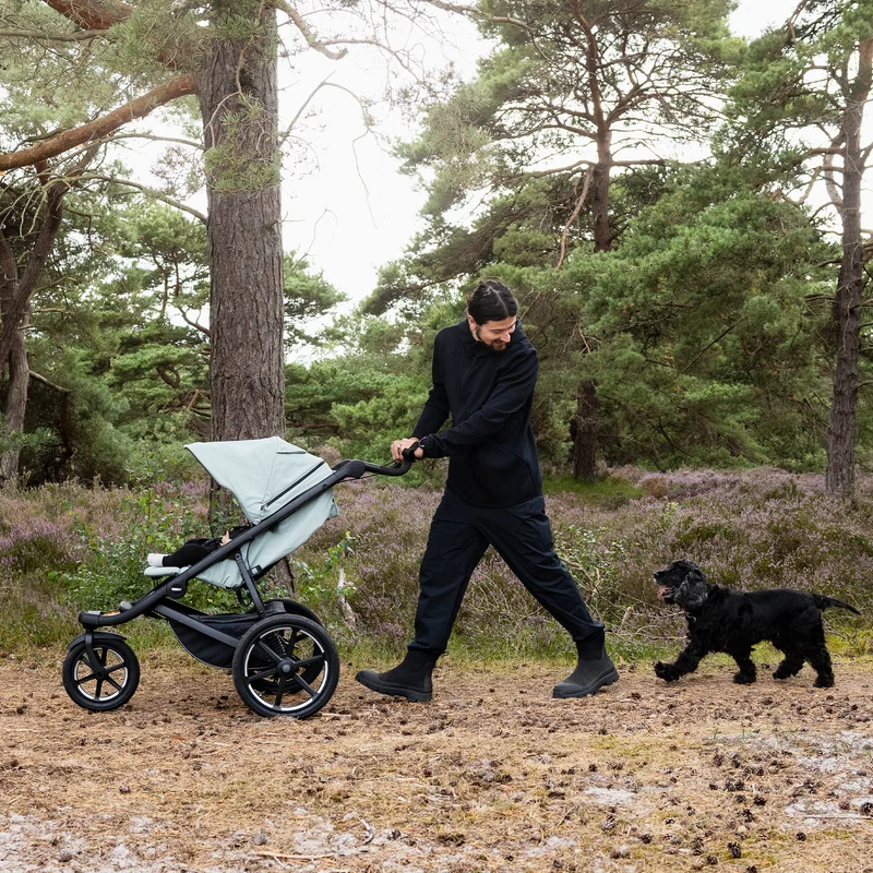 Frau schiebt Thule Urban Glide 3 Kinderwagen mit einem Hund über einen Waldweg.
