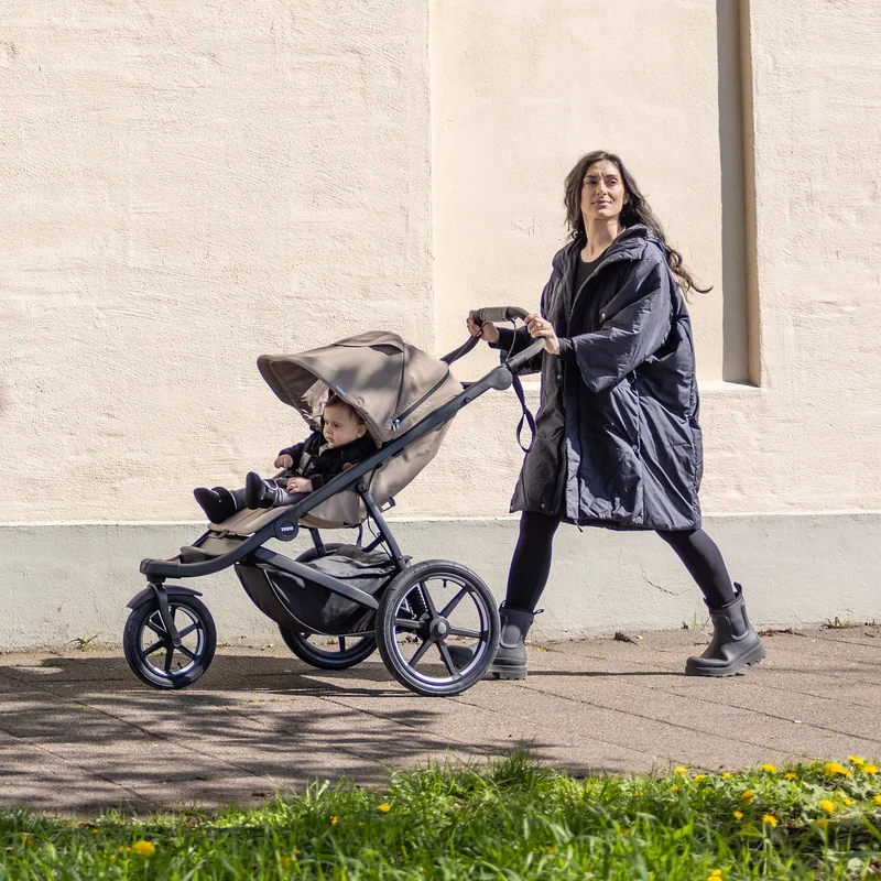 Elternteil hält Kind neben geparktem Thule Urban Glide 3 Kinderwagen auf regnerischer Straße.