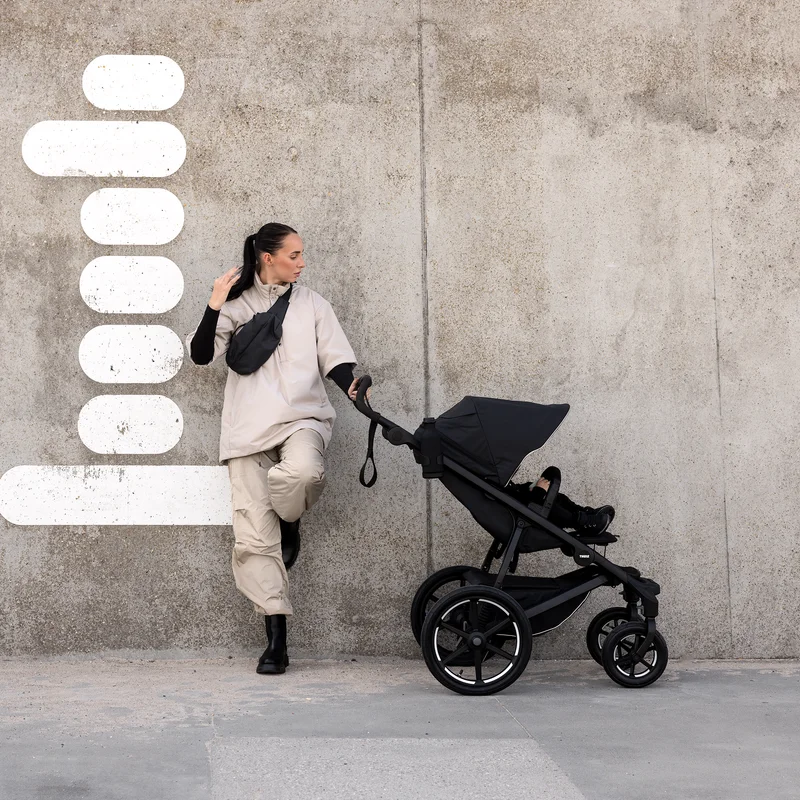 A woman leans against a concrete wall with a black Thule Urban Glide 3 four-wheel all-terrain stroller.
