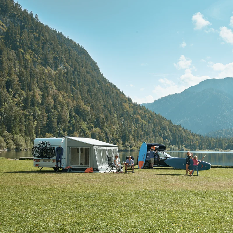 A person walks towards a motorhome with a Thule Veduta rv awning tent, next to a lake and a mountain.